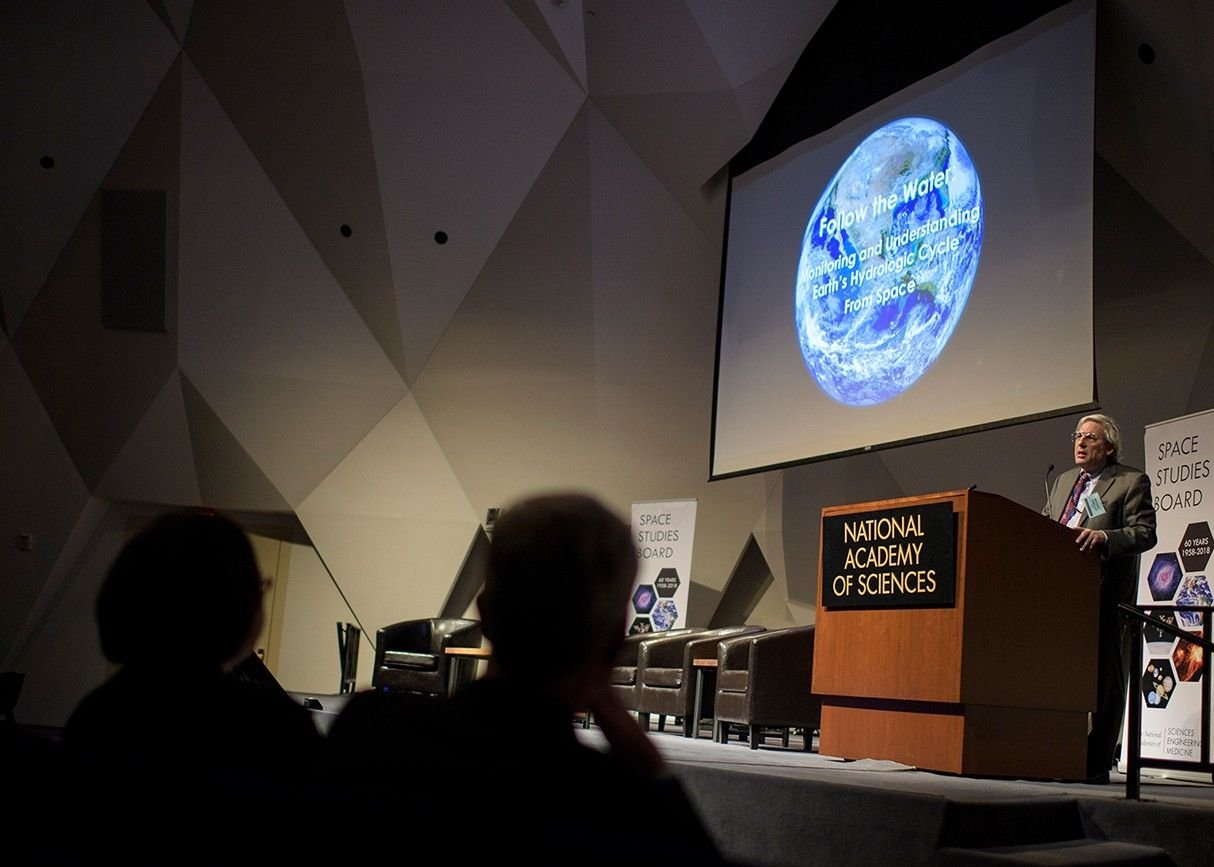 Une conférence scientifique à la National Academy of Sciences, à Washington : la Terre projetée sur grand écran, l’orateur au pupitre, le public en ombre au premier plan.