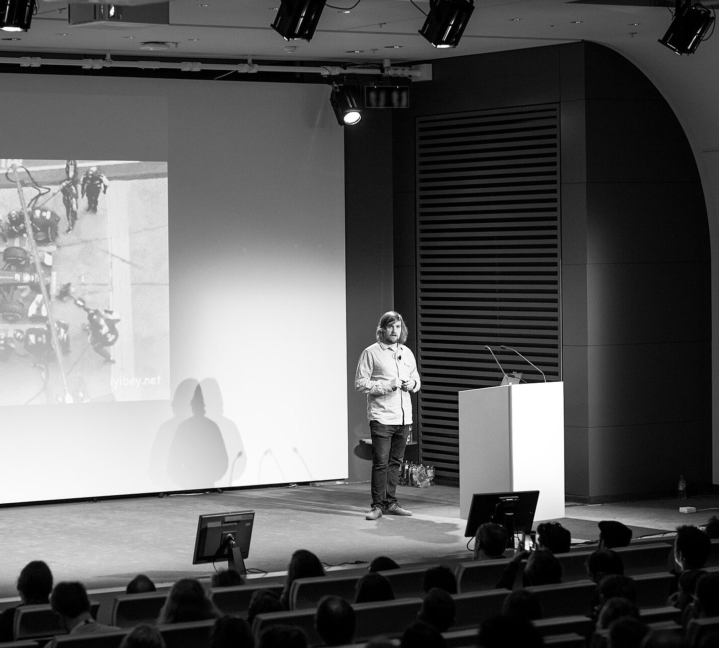 Un conférencier sur une grande scène en 2017, devant l'écran de projection et une salle attentive.